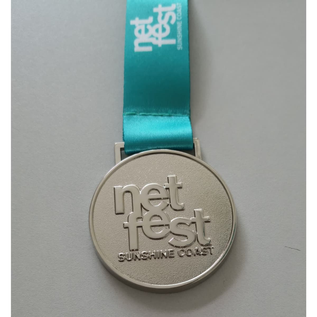 A silver medal with a turquoise ribbon featuring a logo and the text "net fest Sunshine Coast."