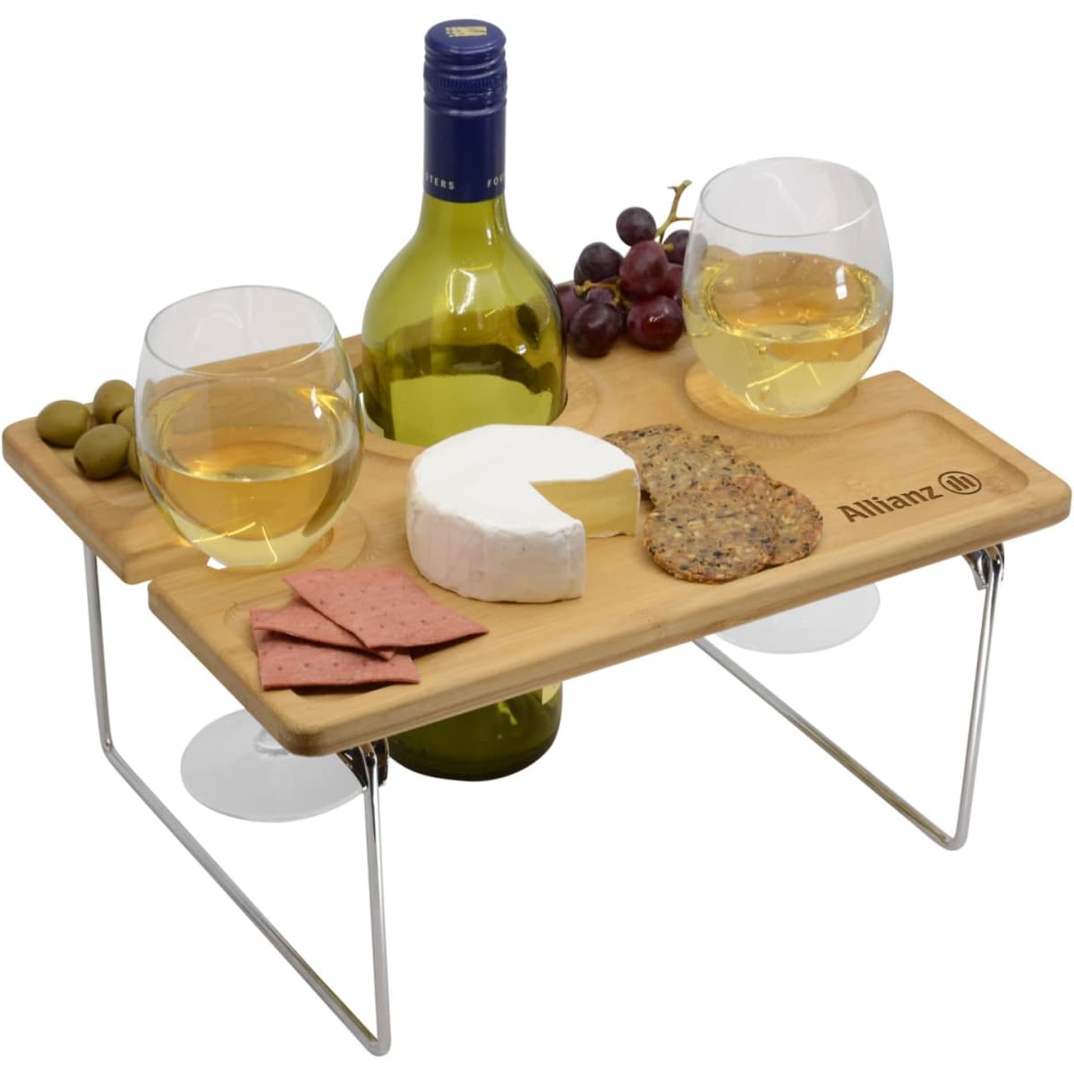 A wooden picnic table with two glasses of white wine, a bottle, cheese, crackers, olives, and grapes.