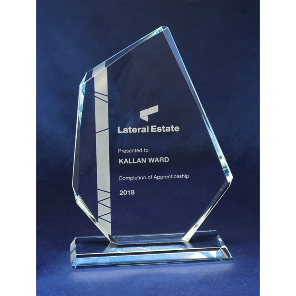 A clear glass award with a unique shape, featuring engraved text and a base, in a blue backdrop.