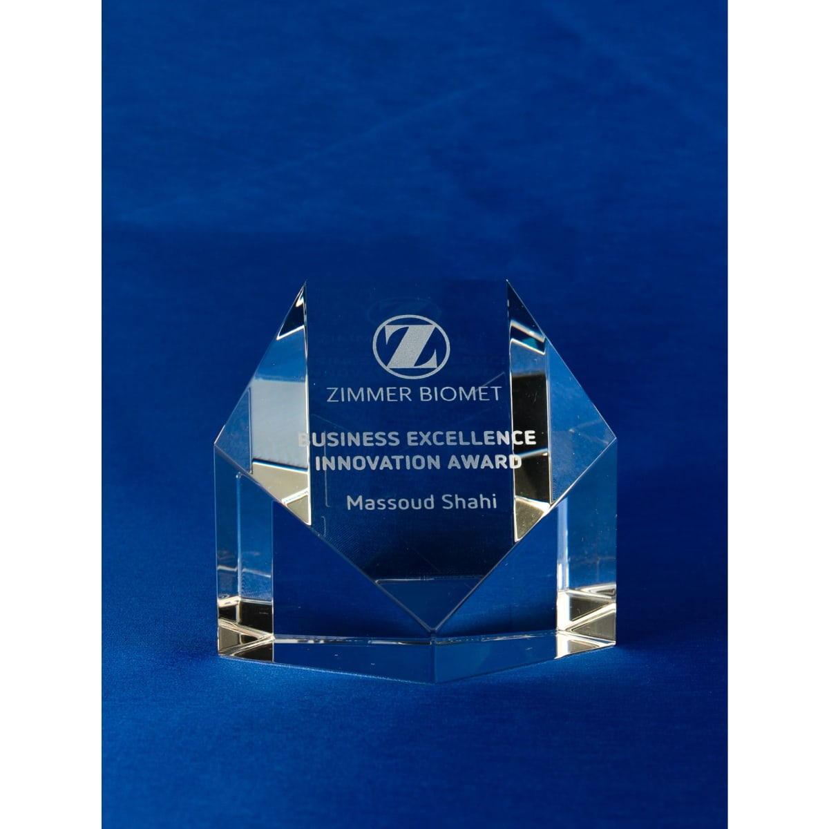 A clear crystal pentagon paperweight with an engraved award, displayed on a blue background. It has a logo and text.