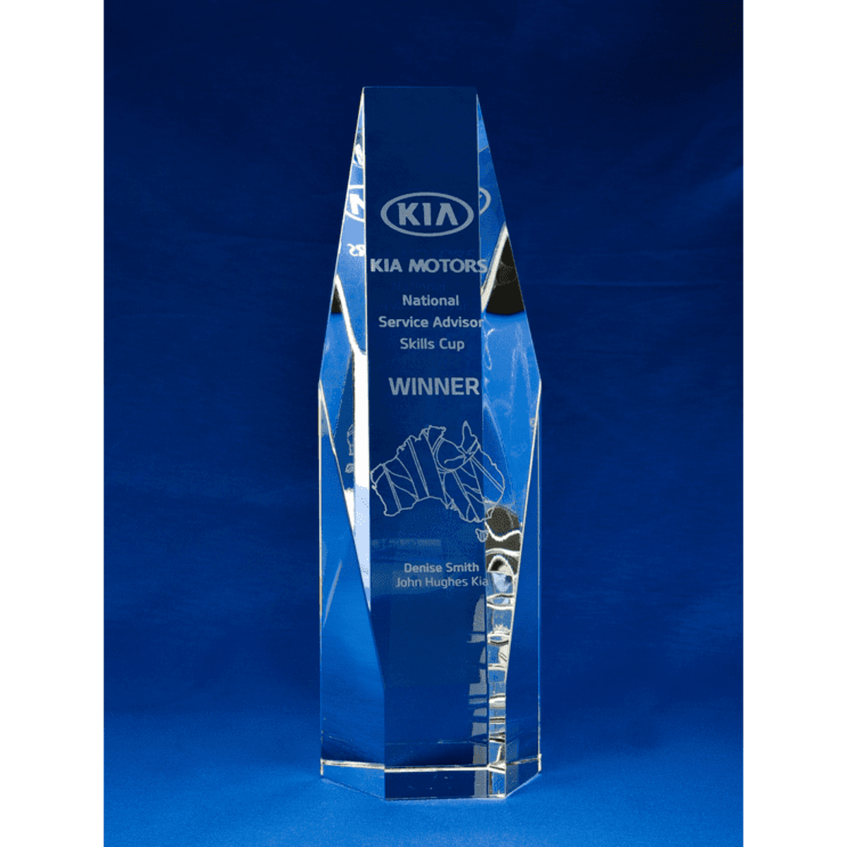 A large, clear crystal trophy in a hexagonal shape, featuring engraving and a logo. The background is blue.