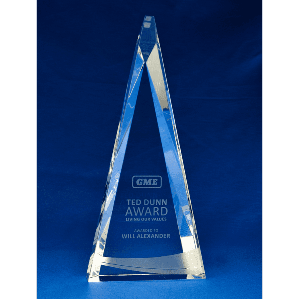 A crystal trophy with a triangular shape, featuring engravings and a logo, set against a blue background.