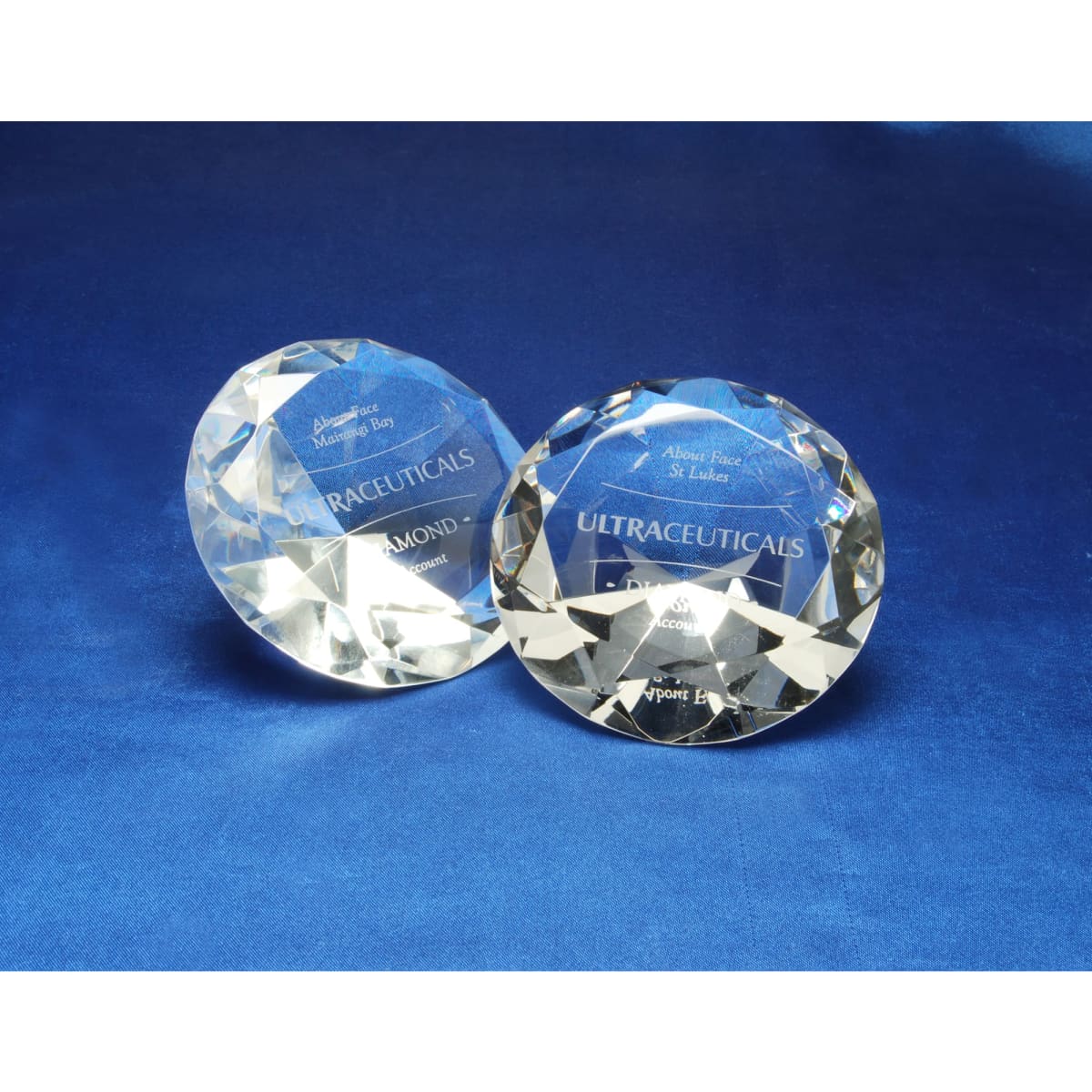 Two crystal diamond awards with a faceted design resting on a blue surface, featuring engraved text and branding.