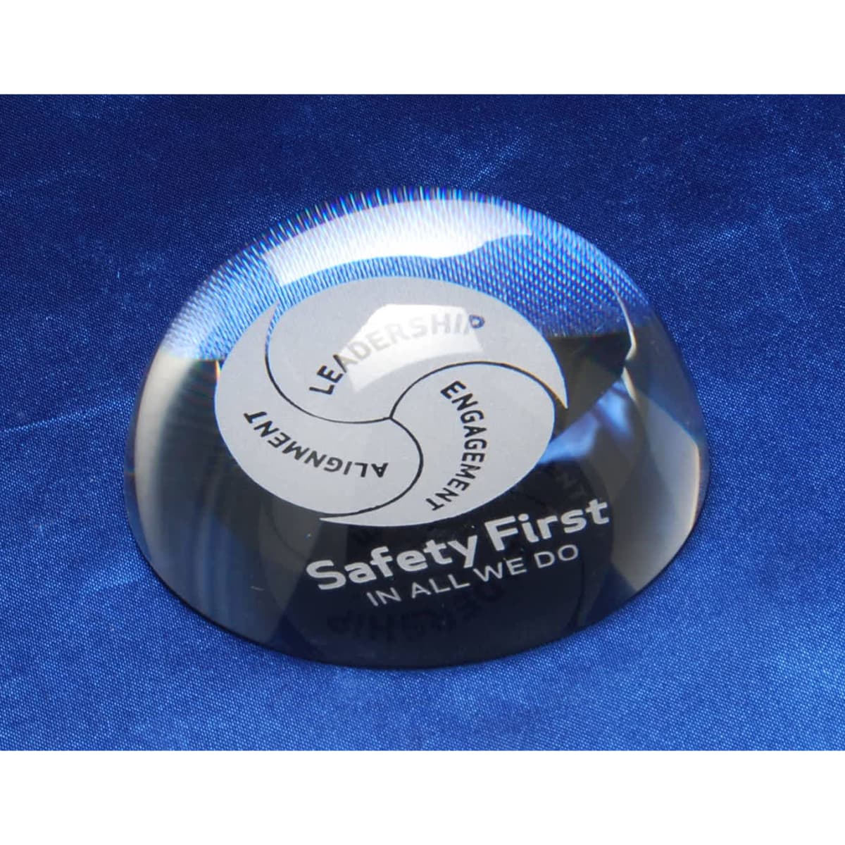 A crystal paperweight with a clear, reflective surface featuring a circular design and a logo. Positioned on a blue fabric background.