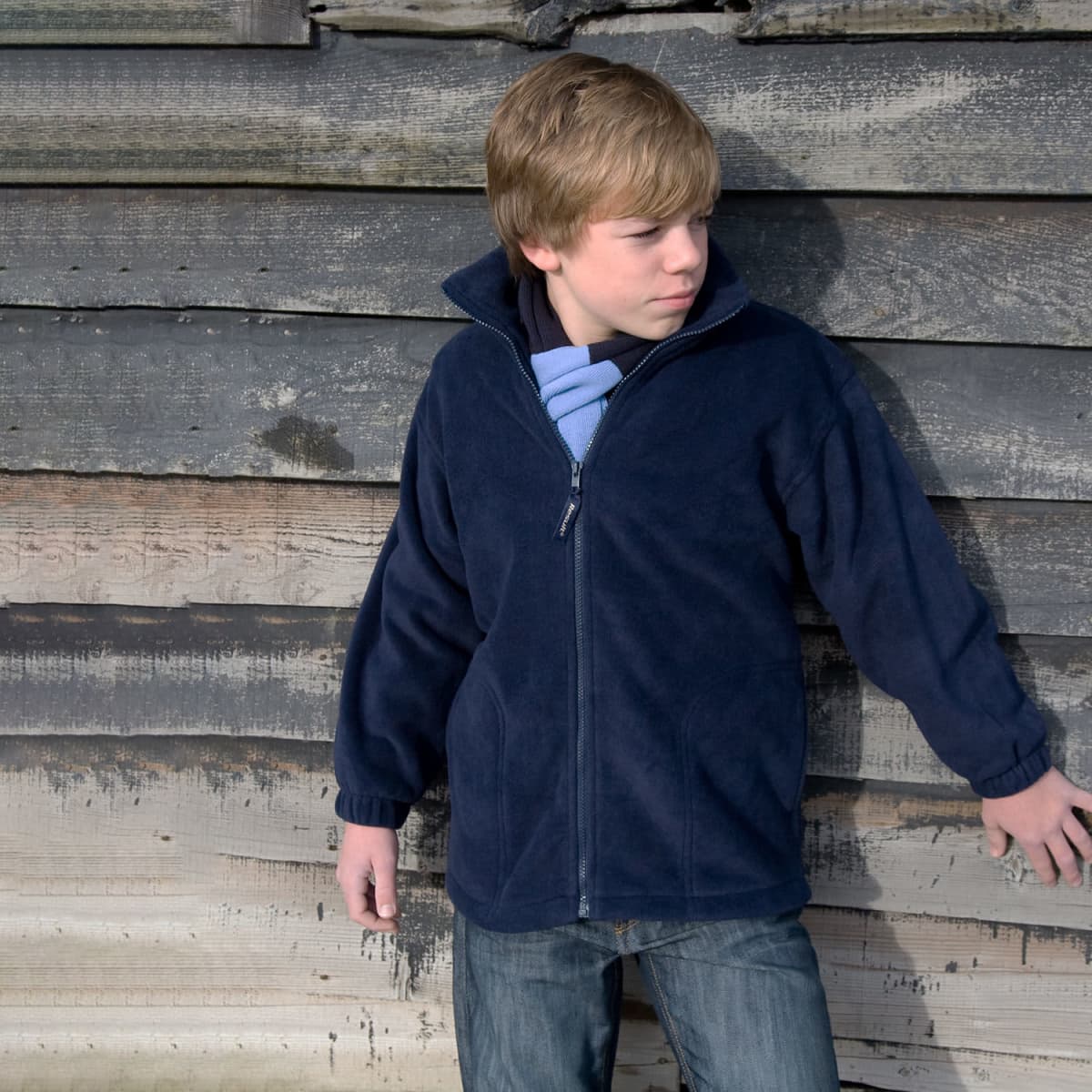 A youth in a navy polar fleece jumper stands against a wooden wall, featuring a full zip and a collar.