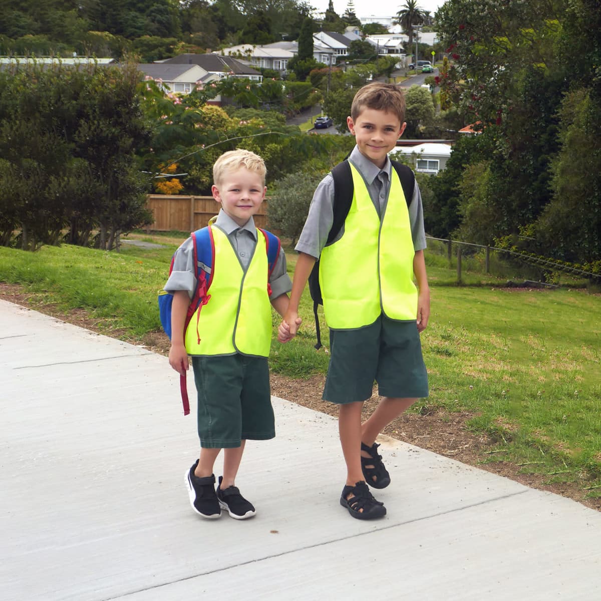 Two children wearing bright yellow safety vests, walking hand in hand on a pathway with greenery and houses in the background.