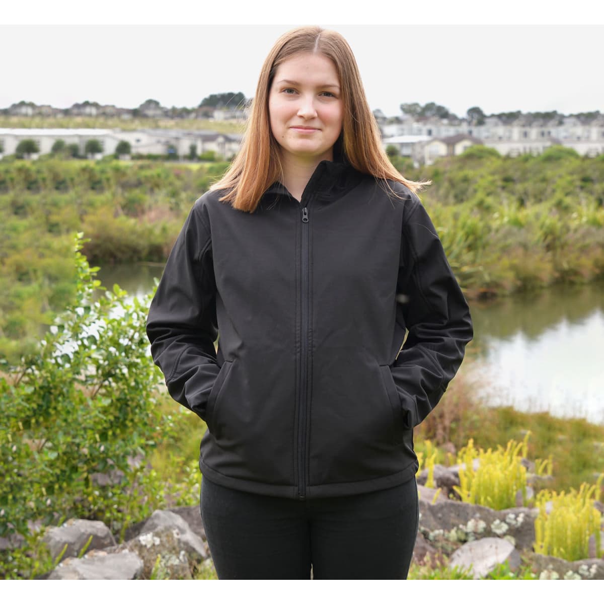 A black softshell jacket features a zip front and side pockets, displayed on a woman outdoors by a serene landscape.