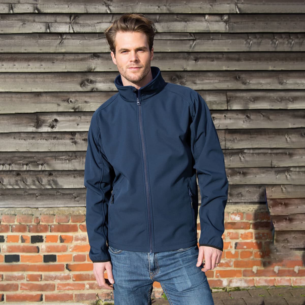 A navy softshell jacket featuring a front zipper and a high collar, modelled by a man against a rustic wooden backdrop.