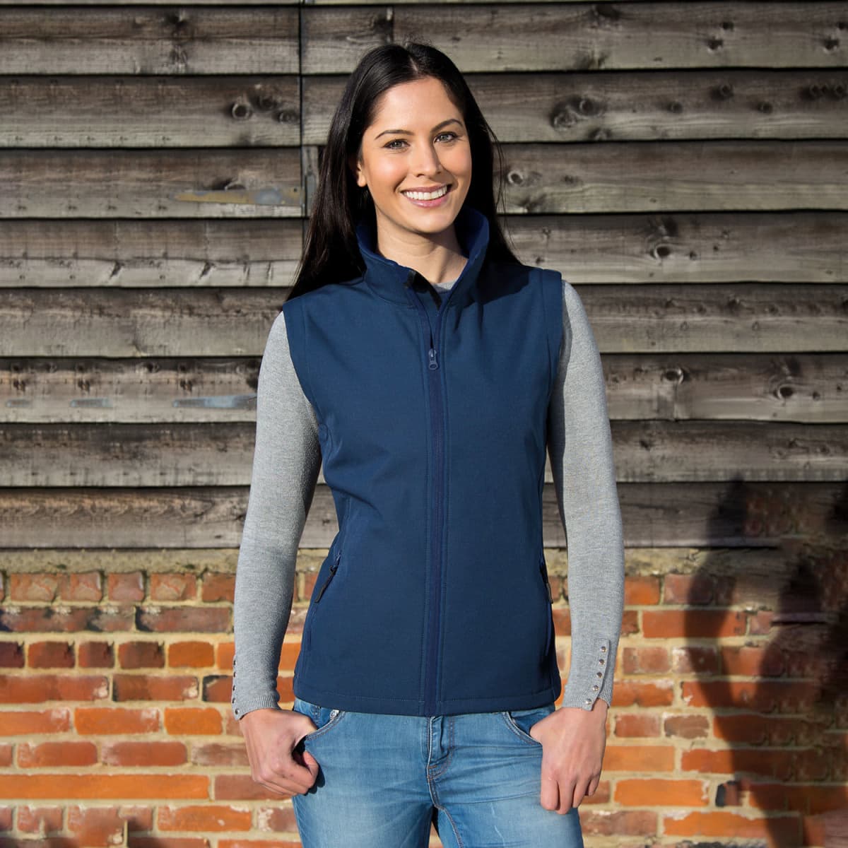 A navy softshell vest worn by a woman, featuring a zip front and pockets, set against a rustic wooden background.