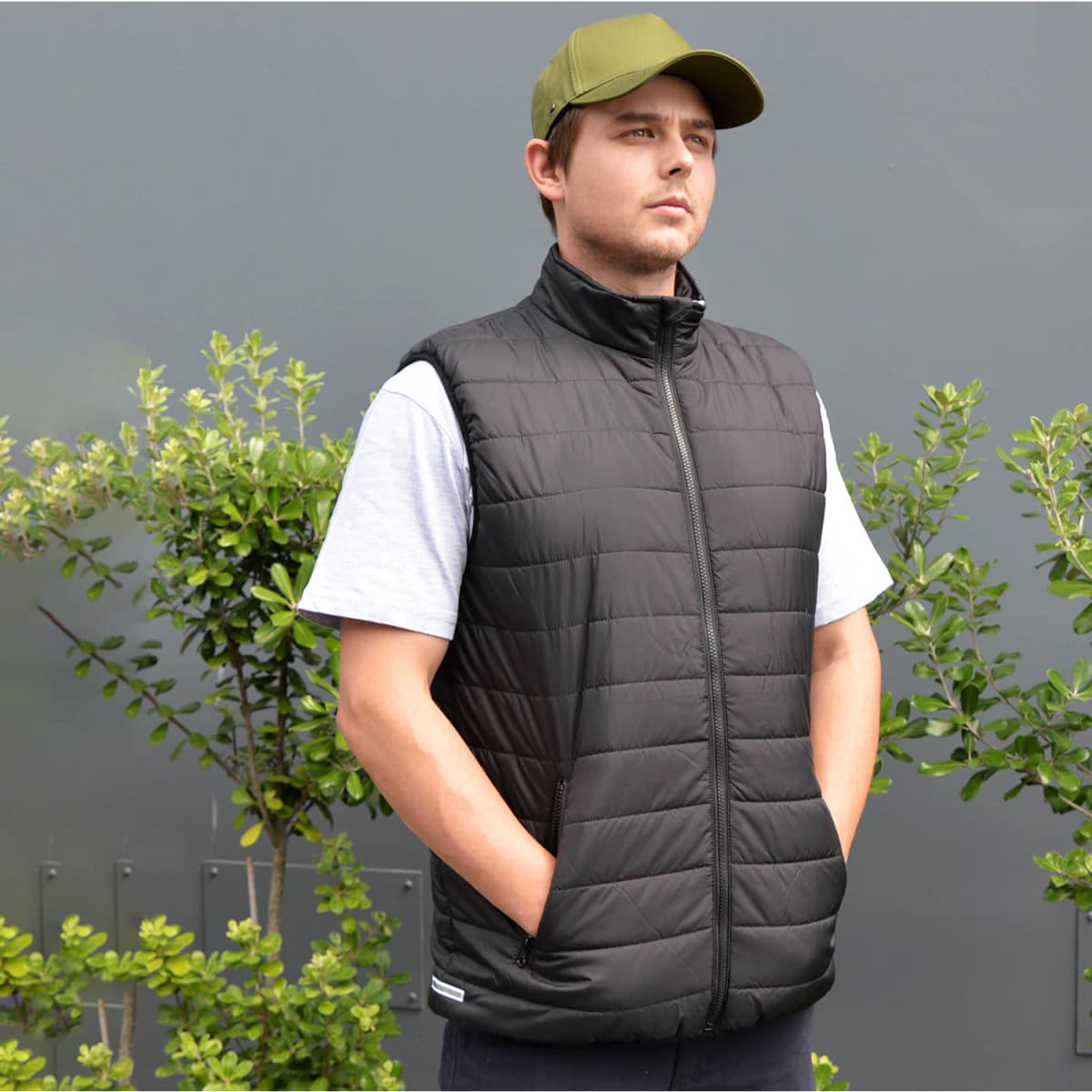 A black padded vest with a high collar worn by a man. The background features green foliage and a grey wall.
