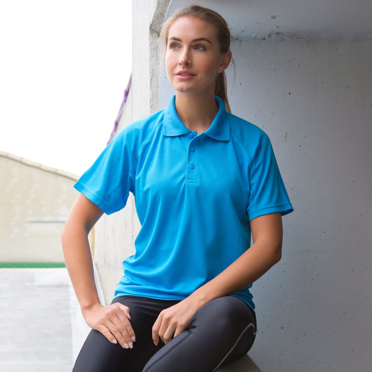 A polo shirt in a vibrant blue colour, featuring a logo, worn by a model seated against a wall.