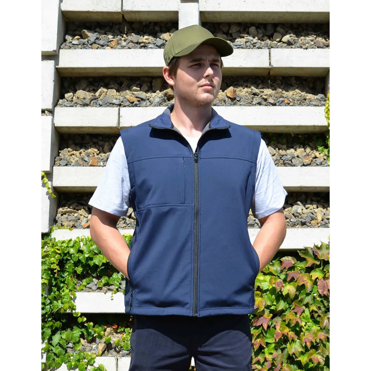 A navy softshell vest with a zip front, pockets, and a cap worn by a man against a stone wall with greenery.