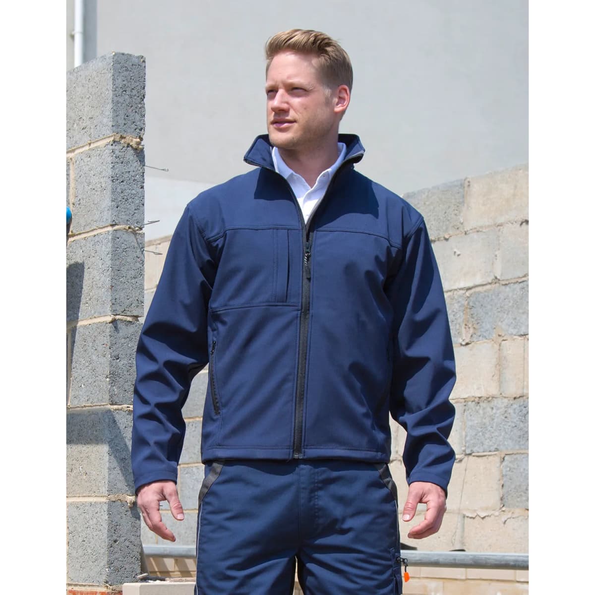 A man wearing a navy soft shell jacket stands against a partially constructed wall, with a branding logo visible.