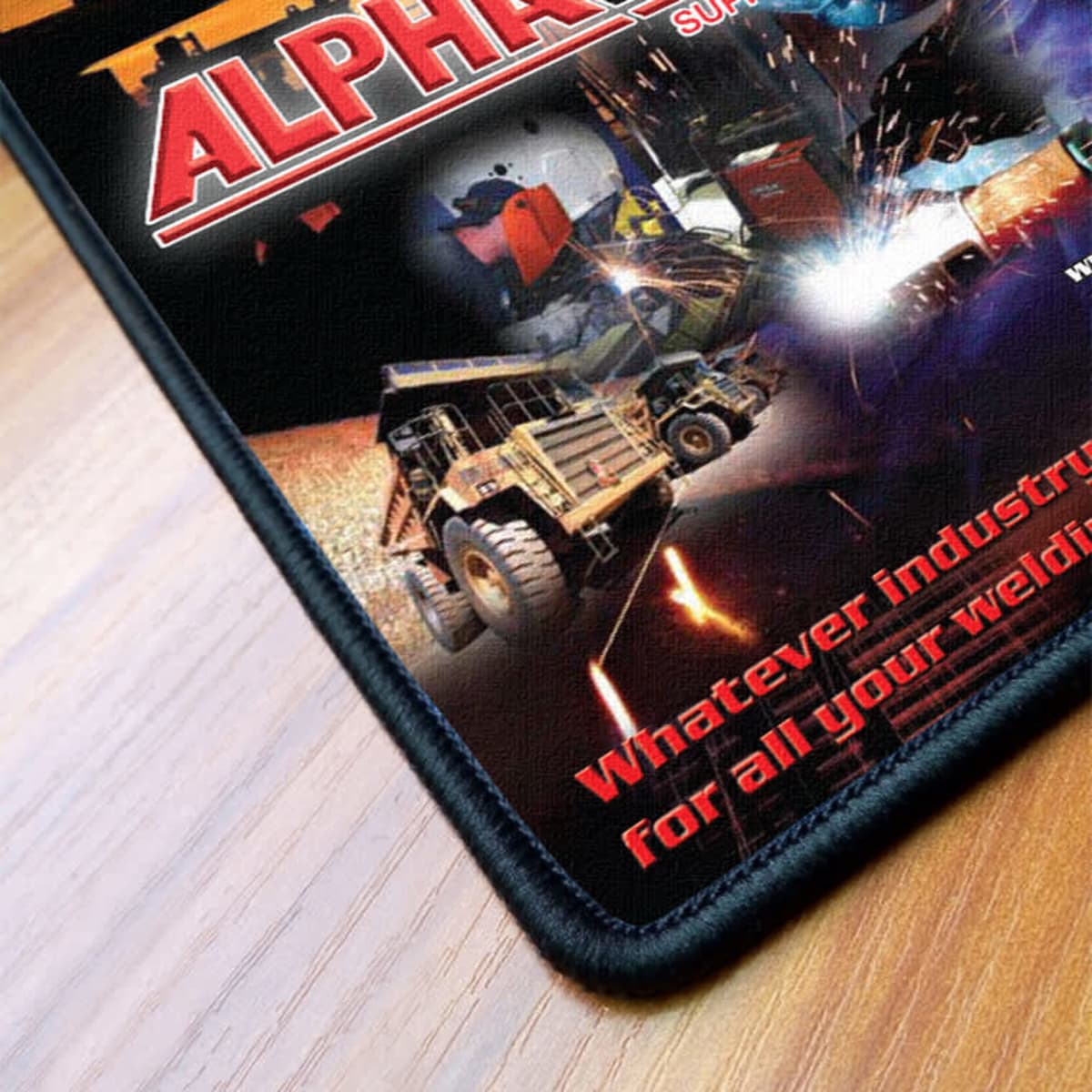 A mousepad featuring a colourful design with industrial vehicles and a logo, bordered with black overlocked fabric.