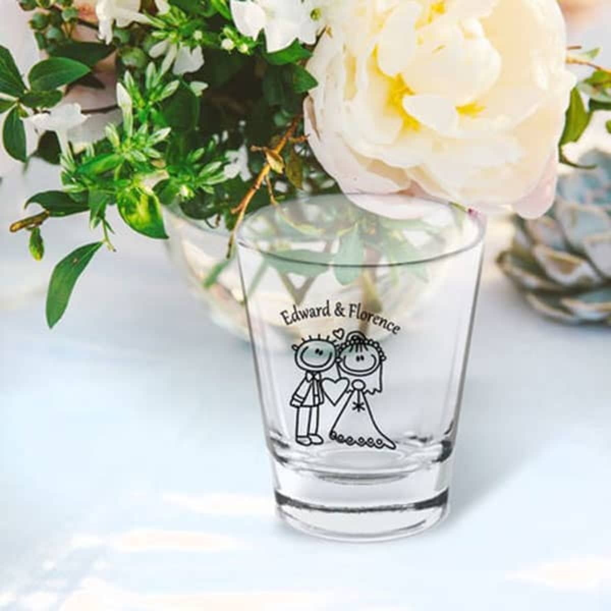 A clear shot glass featuring a couple illustration, surrounded by flowers. The glass has a logo.
