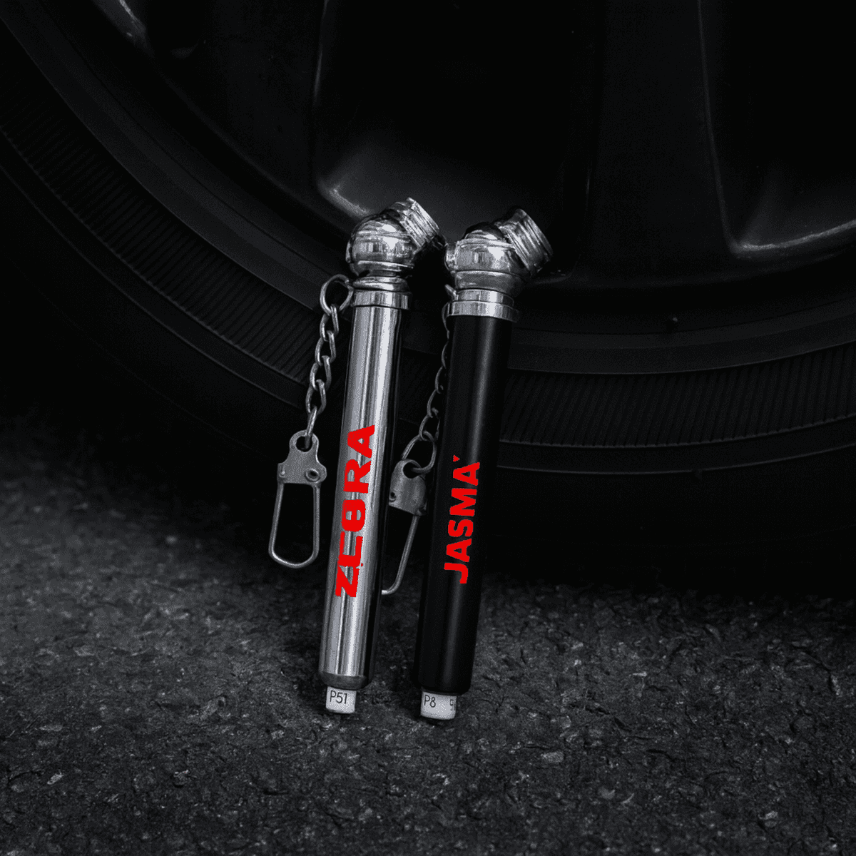 Two key chain tyre gauges, one silver and one black, attached to a keyring, positioned near a tyre.