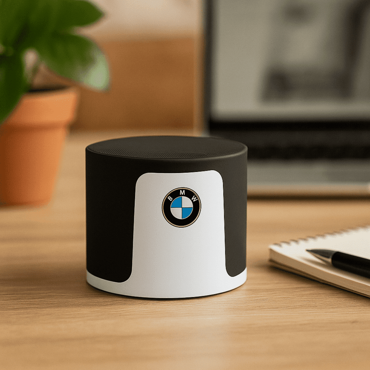 A black and white wireless speaker with a circular design and a logo, placed on a wooden desk near a plant and notebook.