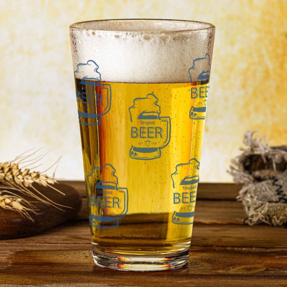 A glass tumbler filled with beer, featuring a decorative pattern and a frothy top, set on a wooden surface.