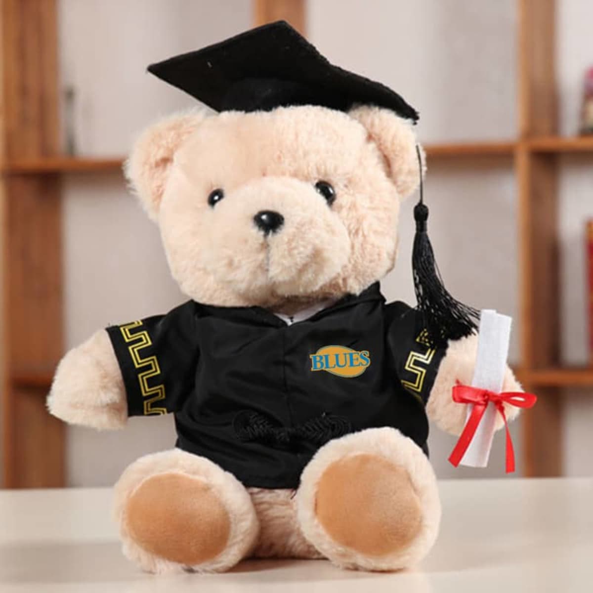 A plush teddy bear dressed in a black graduation gown and cap, holding a rolled diploma with a red ribbon.