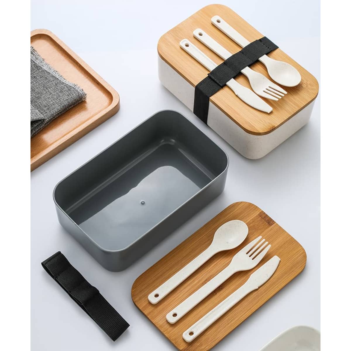 A wheat straw bento lunch box in grey and cream, with bamboo lid and a cutlery set of white utensils.