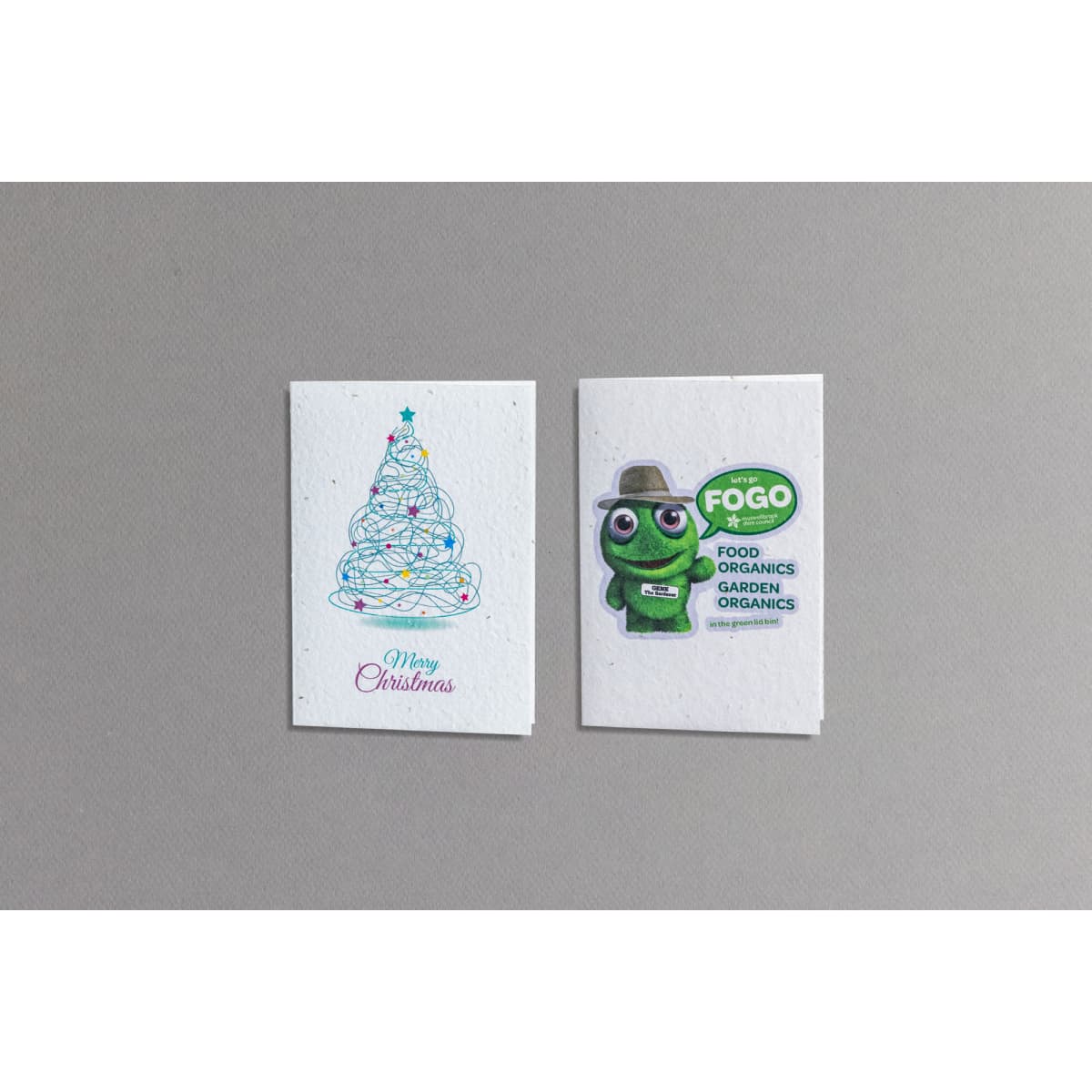 A large seed paper greeting card featuring a Christmas tree design and a green character illustration with a logo.