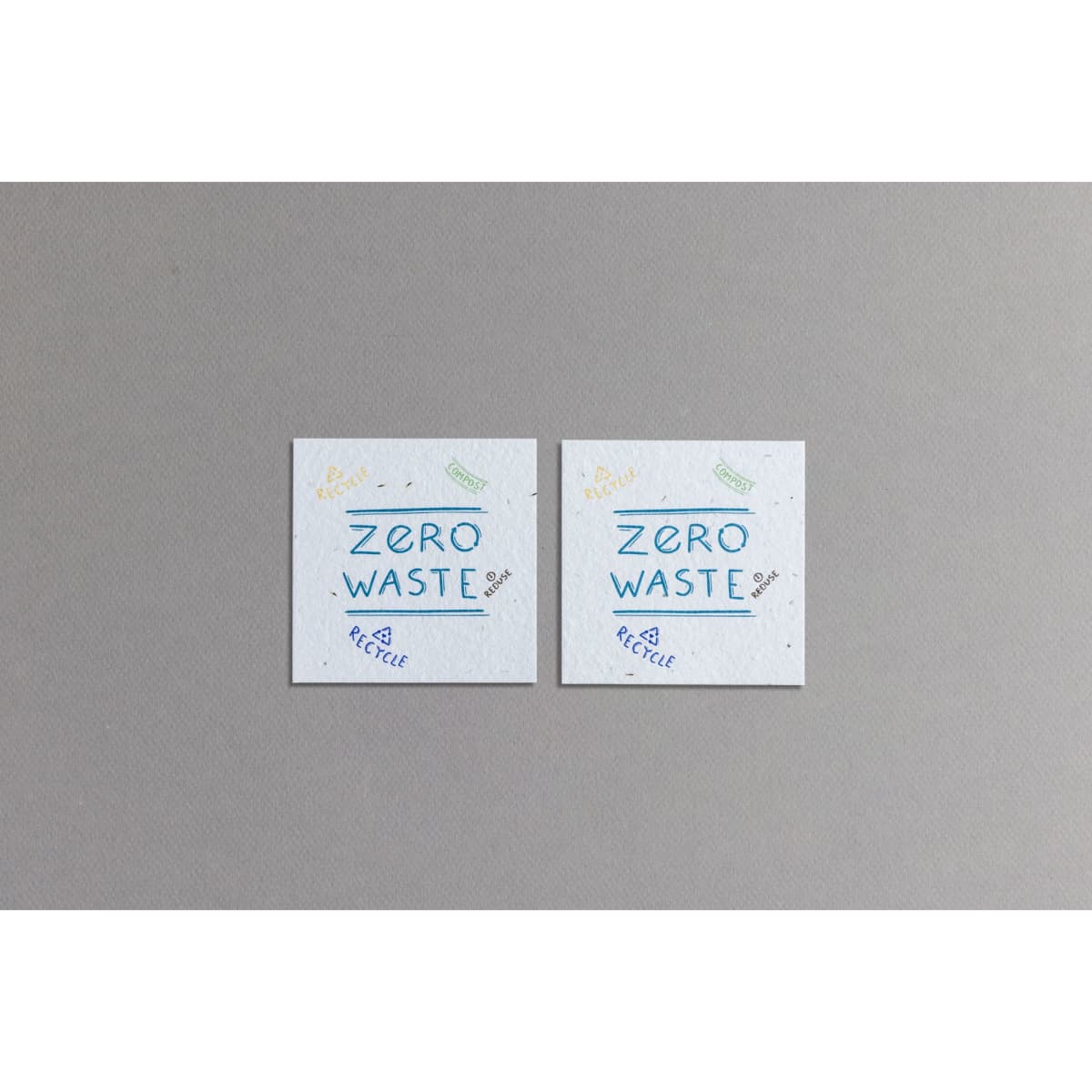 Two square pieces of textured seed paper in light grey with blue and yellow illustrations promoting zero waste.