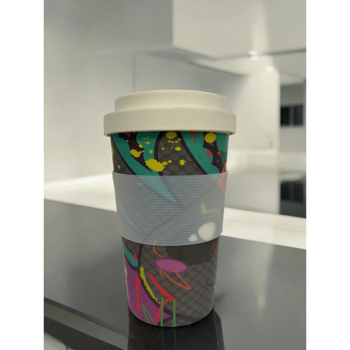 A reusable 400ml coffee cup with a hard lid, featuring a colourful, artistic design and a grey silicone band.