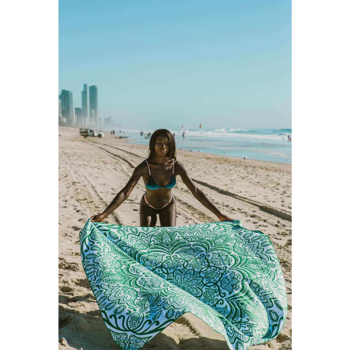 A woman holds a large green and white patterned towel at the beach, with city buildings in the background.