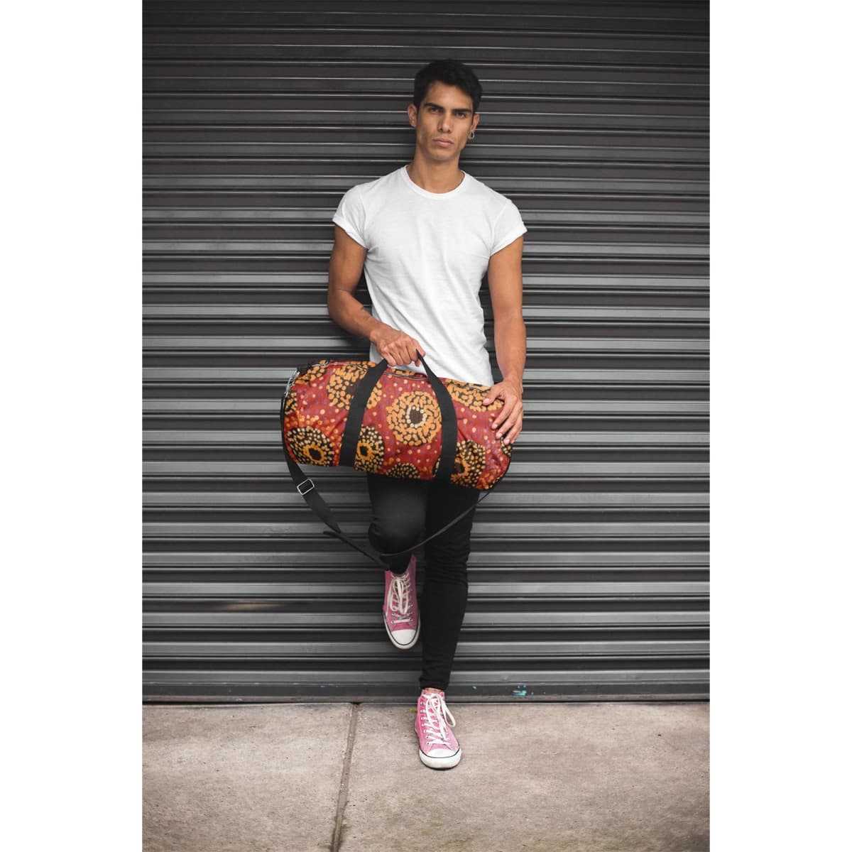 A duffle bag in red and black with a floral pattern is held by a person in a fitted white t-shirt and pink sneakers.