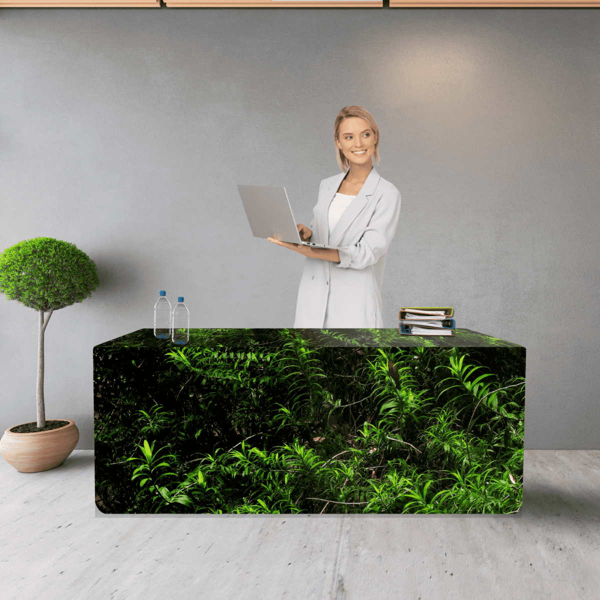 A fitted table cover featuring a green foliage design, with a woman standing behind a table, laptop in hand.