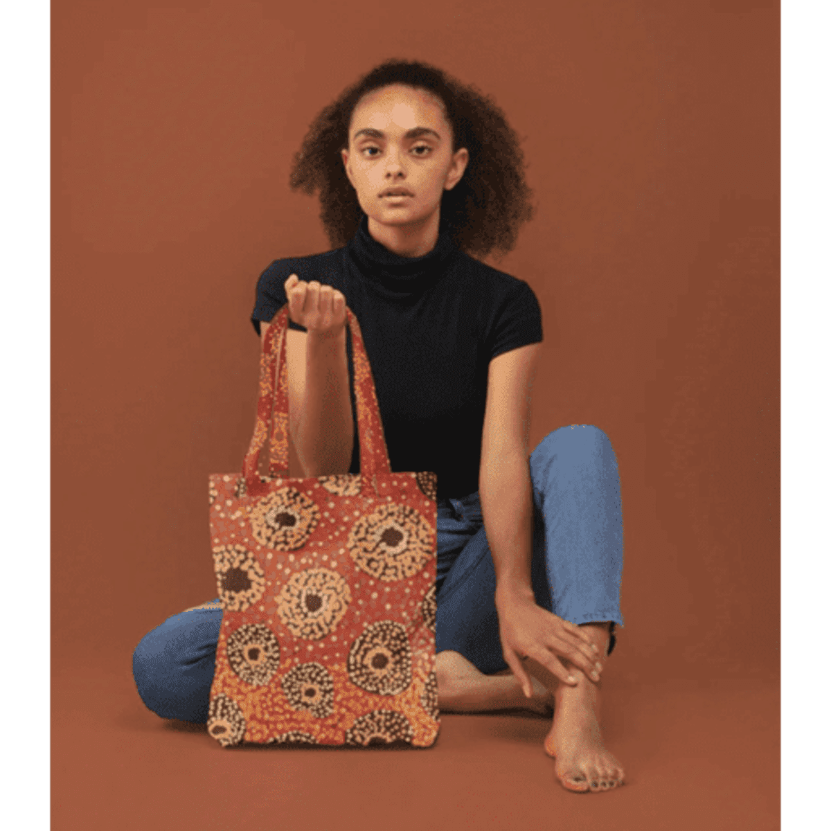 A tote bag featuring a vibrant printed design in red and yellow, held by a model against a brown backdrop.