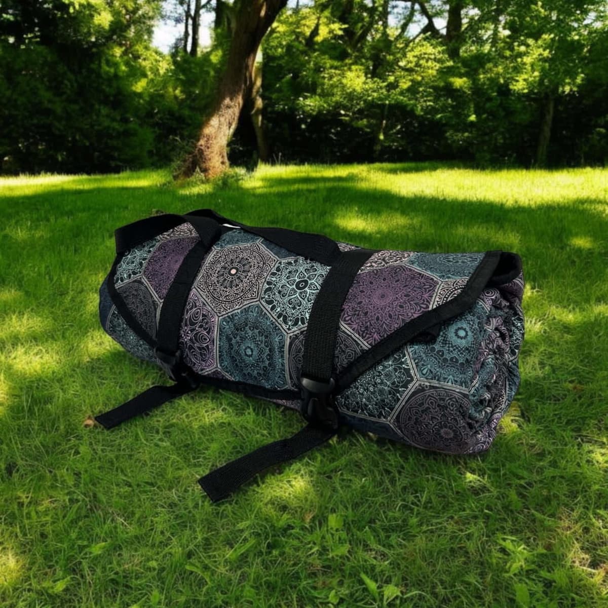 A rolled-up picnic rug in dark purple and teal patterns, secured with straps, resting on green grass.