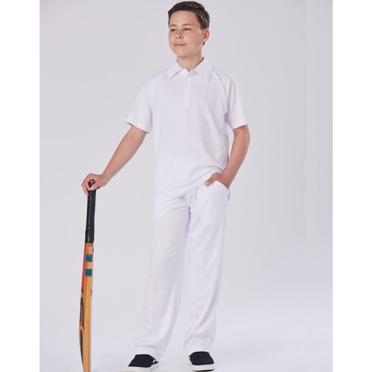 A boy stands confidently wearing white cricket pants and a matching polo shirt, holding a cricket bat.