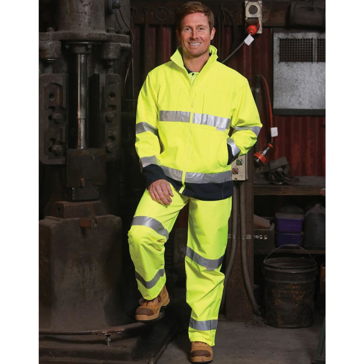 A man stands in a workshop wearing a high-visibility yellow safety pant and jacket with reflective 3M tapes.