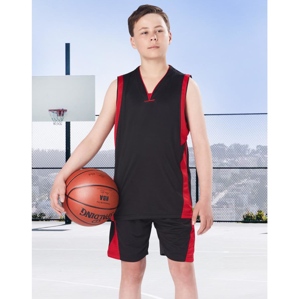 A young boy stands on a basketball court wearing black and red basketball shorts and a matching sleeveless top, holding a basketball.
