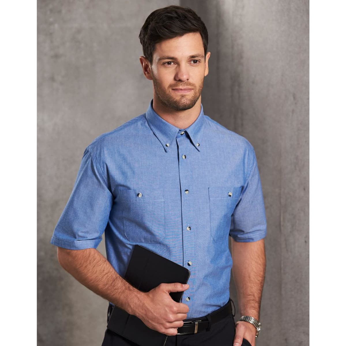 A man wearing a short sleeve chambray shirt in light blue, holding a folder. The shirt features a classic button-up design.