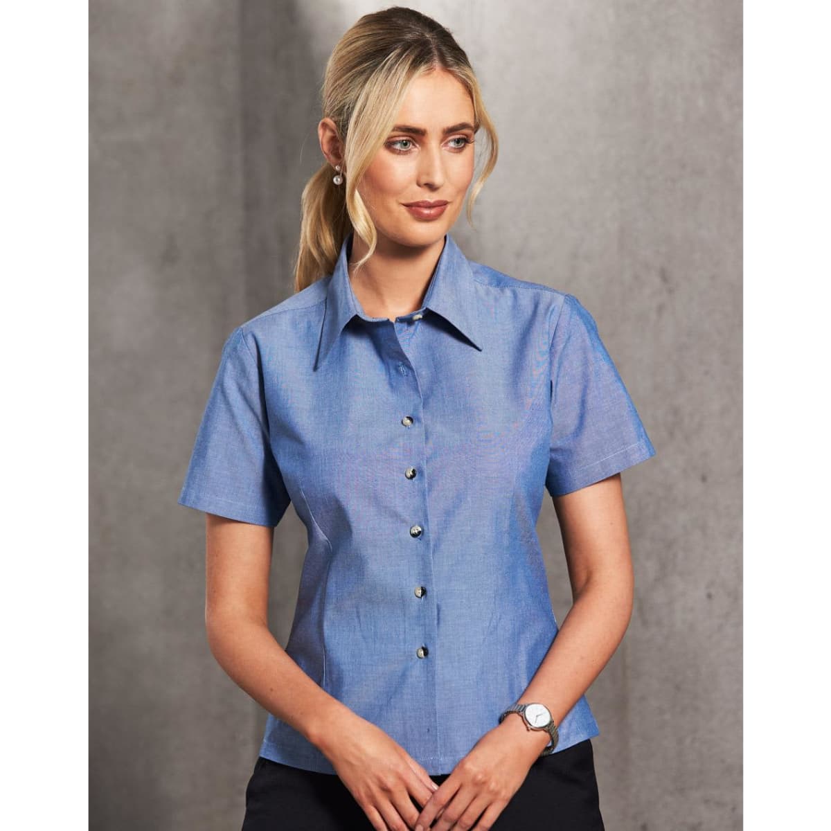 A button-up ladies' short sleeve shirt in chambray blue, featuring a collar and a row of buttons, with a logo.