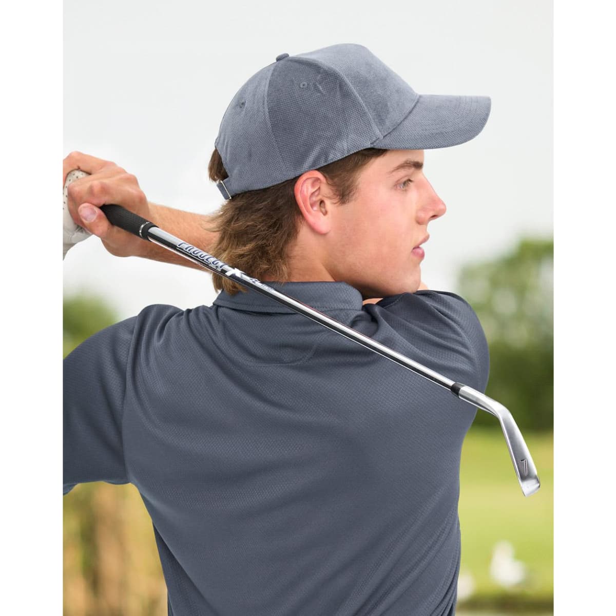 A young man wearing a grey jacquard texture 5-panel cap, holding a golf club in a lush outdoor setting.