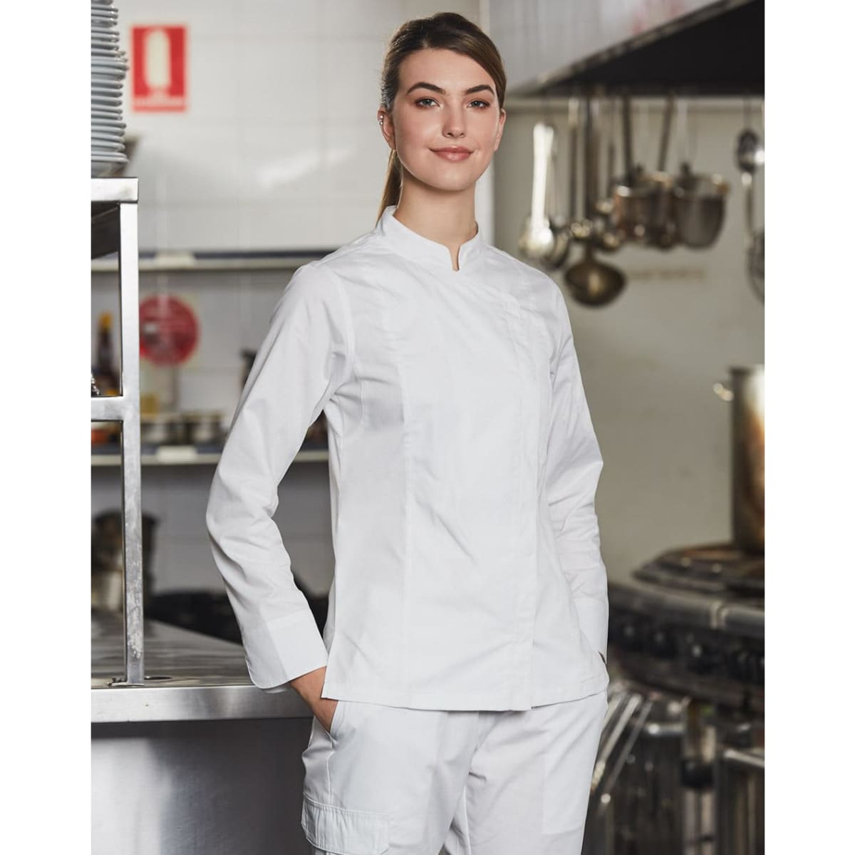 A white ladies' functional chef jacket displayed on a model in a professional kitchen setting. The jacket has a logo.