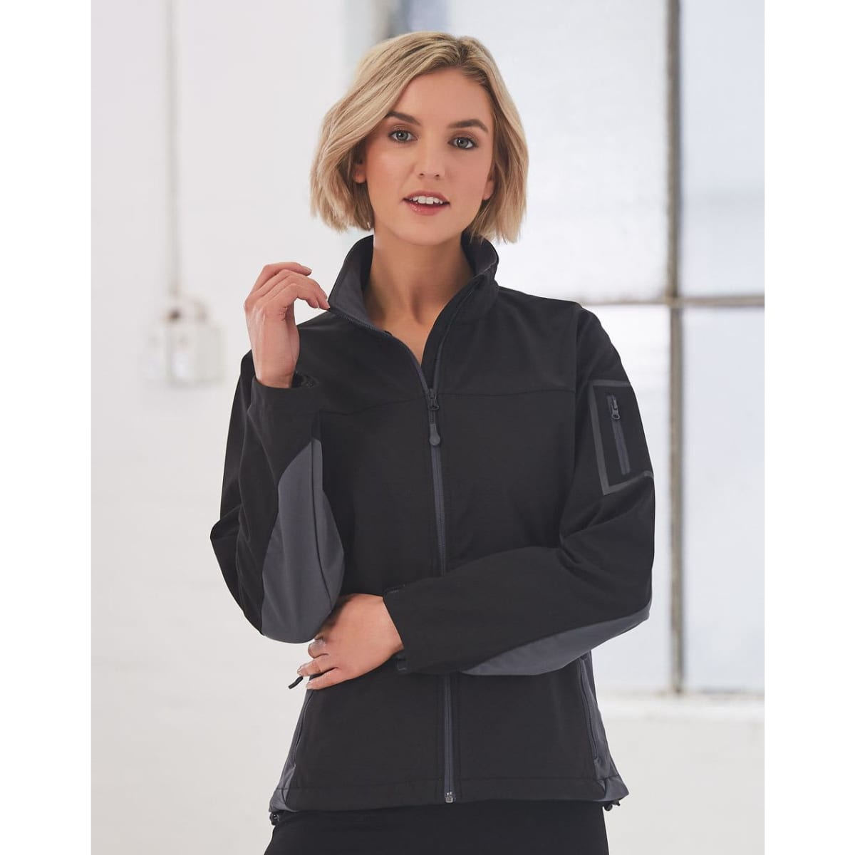 A black softshell jacket with grey accents, featuring a zip front and a side pocket, worn by a model indoors.
