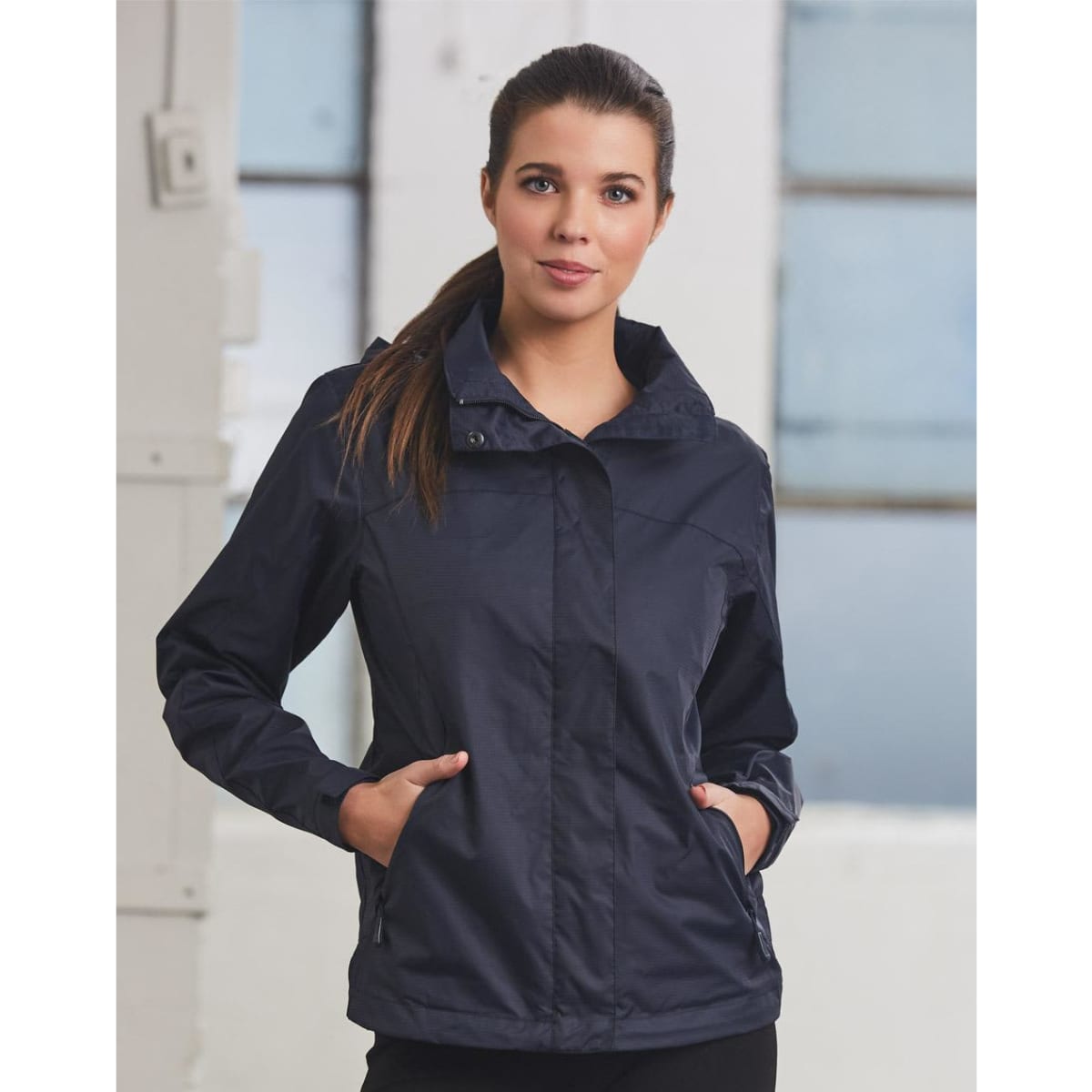 A navy versatile jacket featuring a hood and side pockets, modelled by a woman against an industrial backdrop.