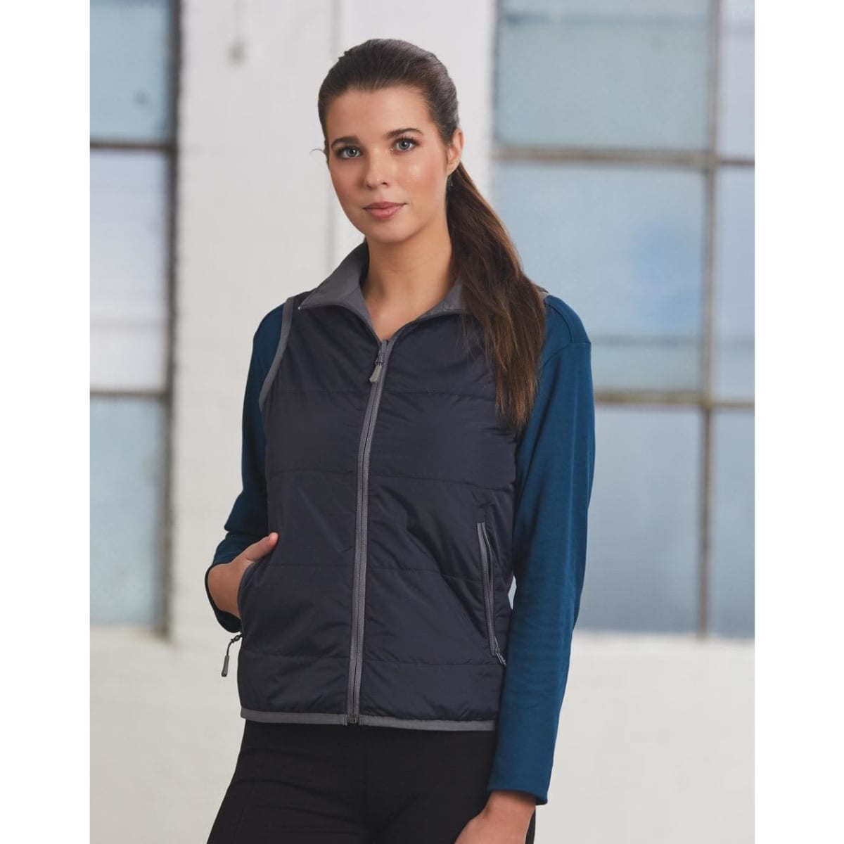 A woman models a navy blue versatile vest with grey accents over a long-sleeve top, featuring pockets and a zip.