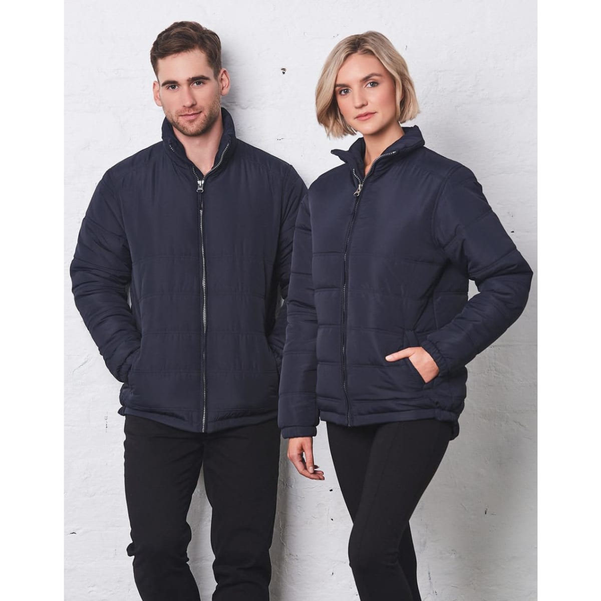 A navy puffer jacket is worn by a man and woman, standing side by side against a light backdrop.