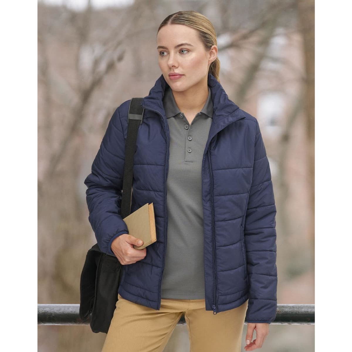 A woman in a navy puffer jacket stands outdoors, holding a book and wearing a grey polo shirt and tan trousers.