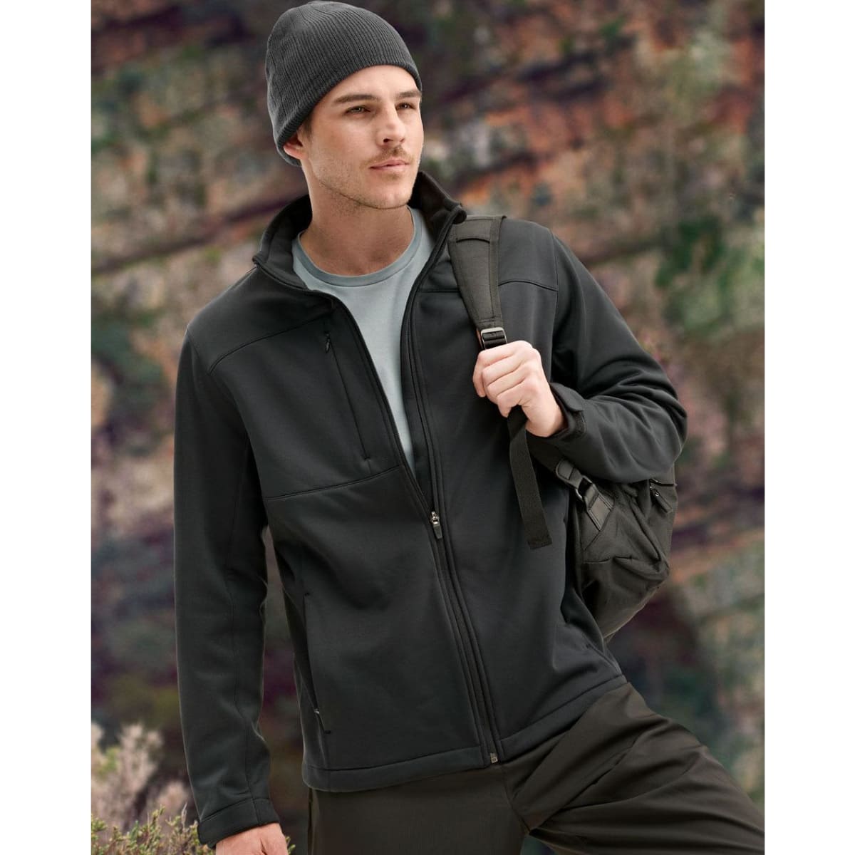 A men's sustainable softshell jacket in black, featuring a zip front and a logo, worn with a beanie and backpack.