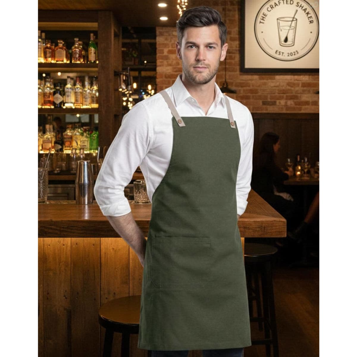 A person wearing a light brown canvas bib apron over a grey t-shirt, standing against a brick wall.