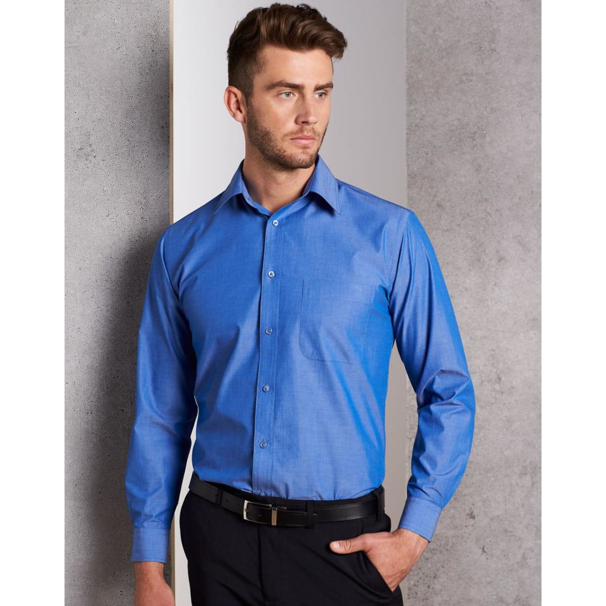 A blue long sleeve button-up shirt with a chest pocket and a logo, worn by a man against a grey background.