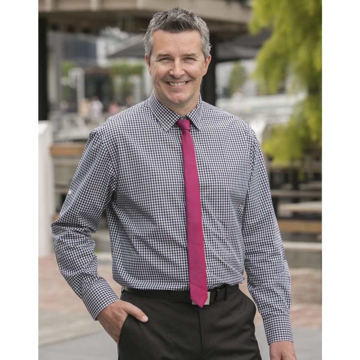 A man in a blue and white gingham check long-sleeve button-up shirt, wearing a pink tie and black trousers.