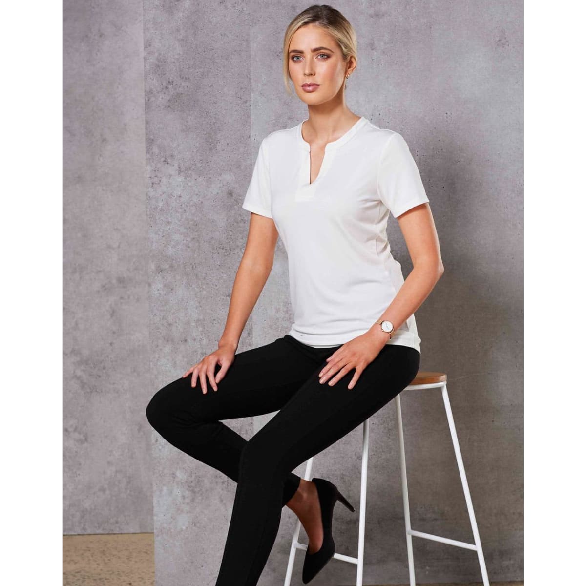A white ladies' V-neck short-sleeve knit top is worn with black pants, featuring a logo. Model seated on a bar stool.