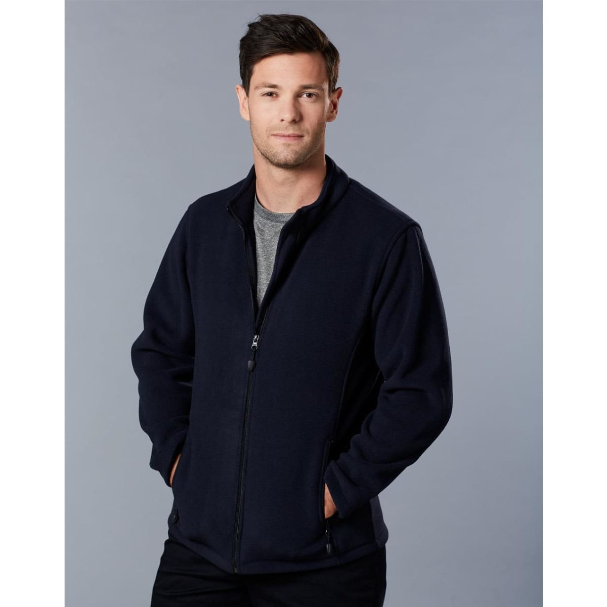 A man in a navy polar fleece jacket with a full zip, standing with hands in pockets against a grey background.